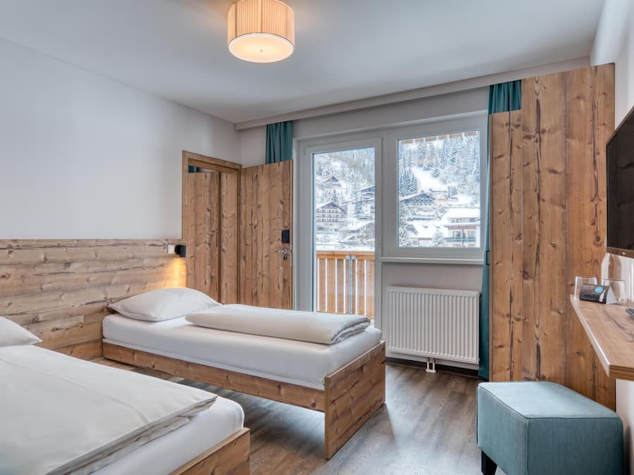 Ein Zimmer mit zwei Betten, Holzwänden, einem Fenster mit Blick auf schneebedeckte Gebäude und einem Schreibtisch mit Stuhl.