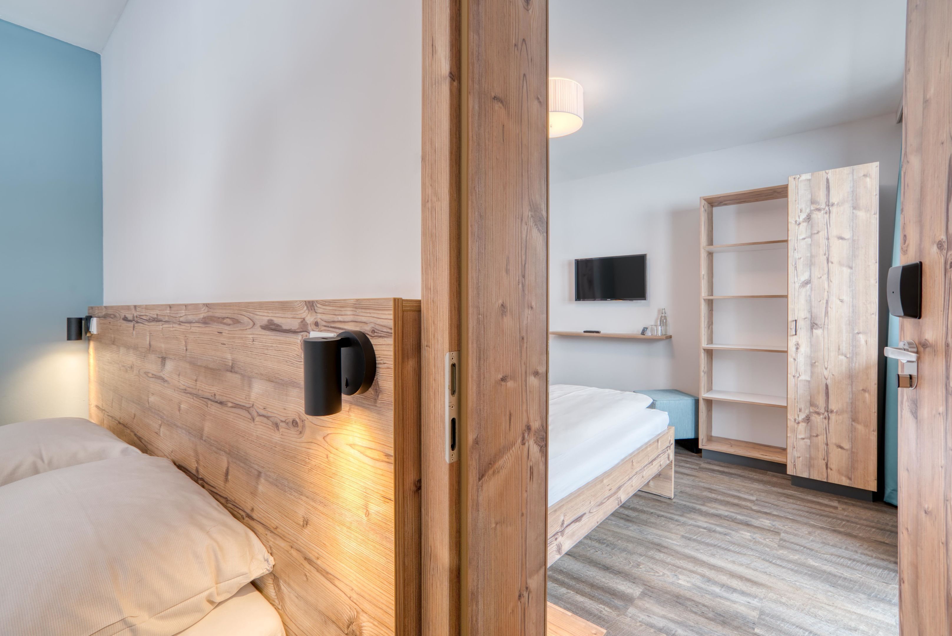 Moderni hotellihuone, jossa on puinen sängynpääty, sänky, seinälle asennettu televisio ja sisäänrakennettu puinen vaatekaappi.