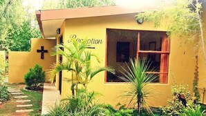 A reception area with a cross on the wall, a window with red curtains, and a pathway leading to the entrance. - At Home Guesthouse (Upington)