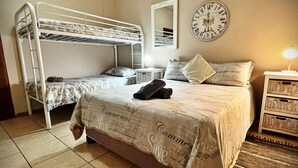 A room with bunk beds, a bedside table, a lamp, and a wall clock. - At Home Guesthouse (Upington)