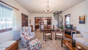 Una sala de estar con sofá, un sillón estampado, una mesa de comedor con sillas, un mueble para la televisión y una escalera en espiral.