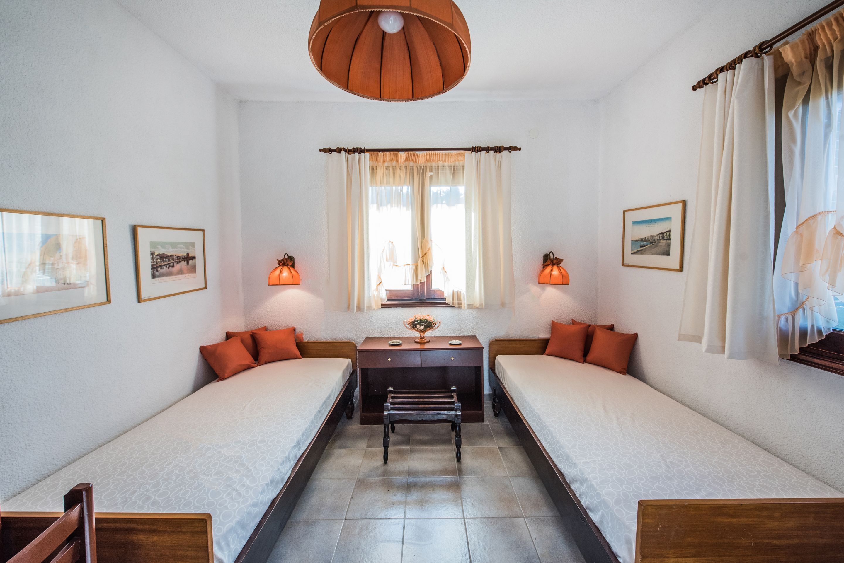 A room with two single beds, a wooden bench, a small table with a vase, and framed pictures on the walls.