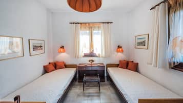 A room with two single beds, a wooden bench, a small table with a vase, and framed pictures on the walls.