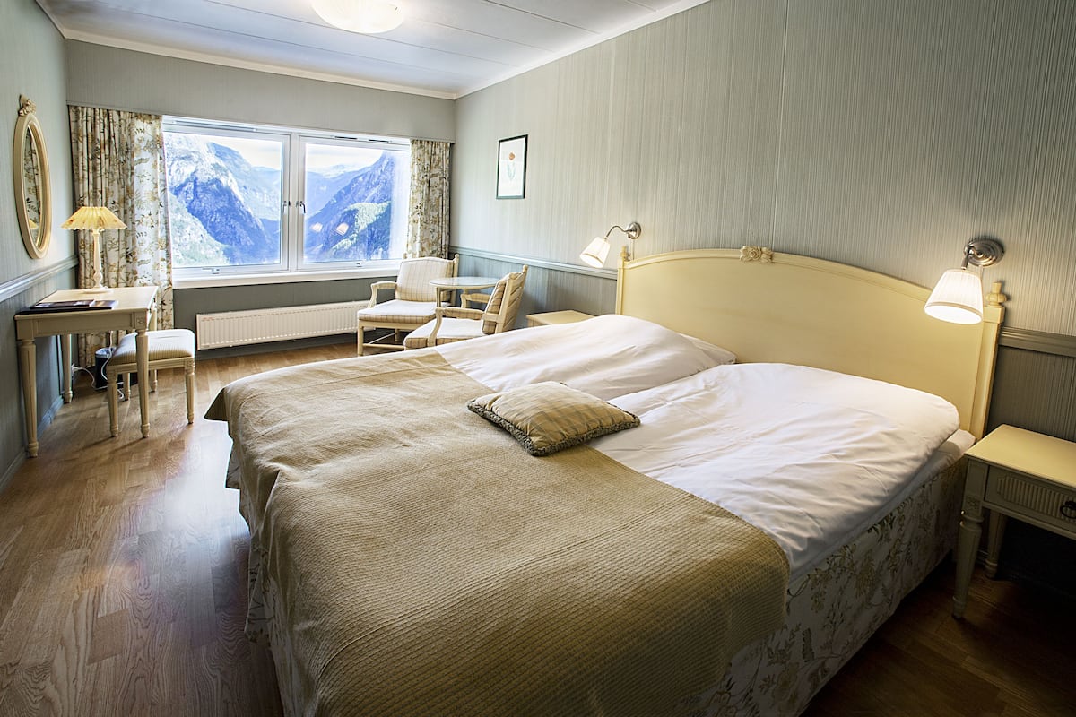 A hotel room with a large bed, a desk with a lamp, a chair, and a view of mountains through the window.