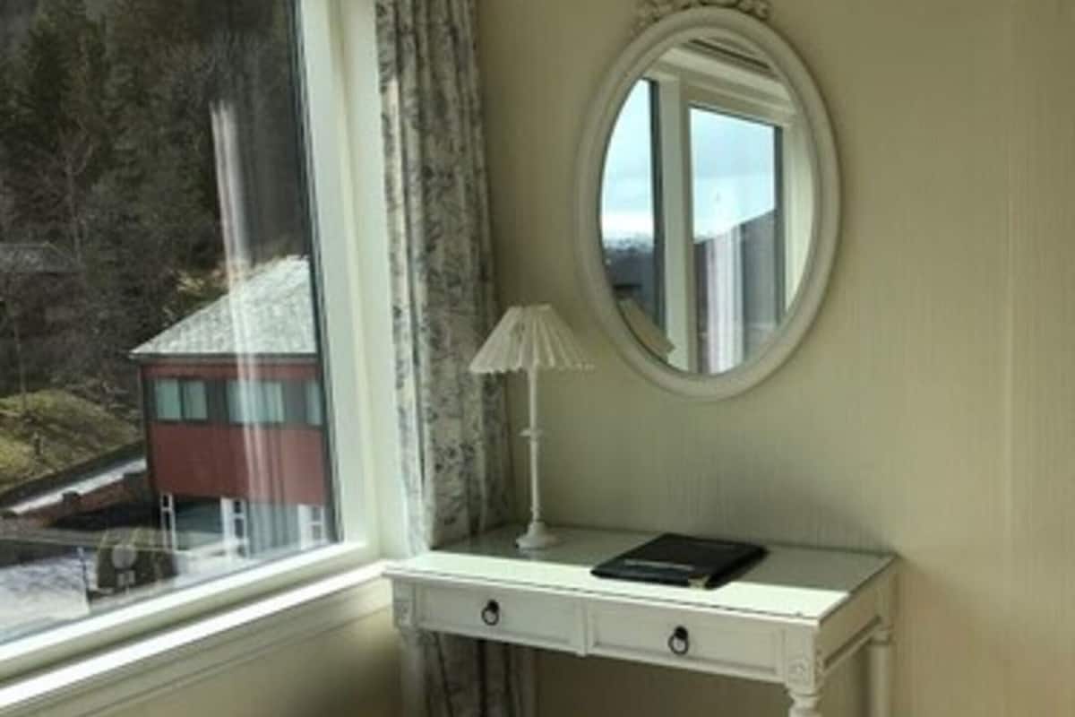 A room with a white desk, a round mirror, a lamp, and a window with curtains.
