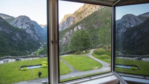 Vista a través de una ventana que muestra un paisaje montañoso con una zona de césped y un camino que lleva a un edificio.