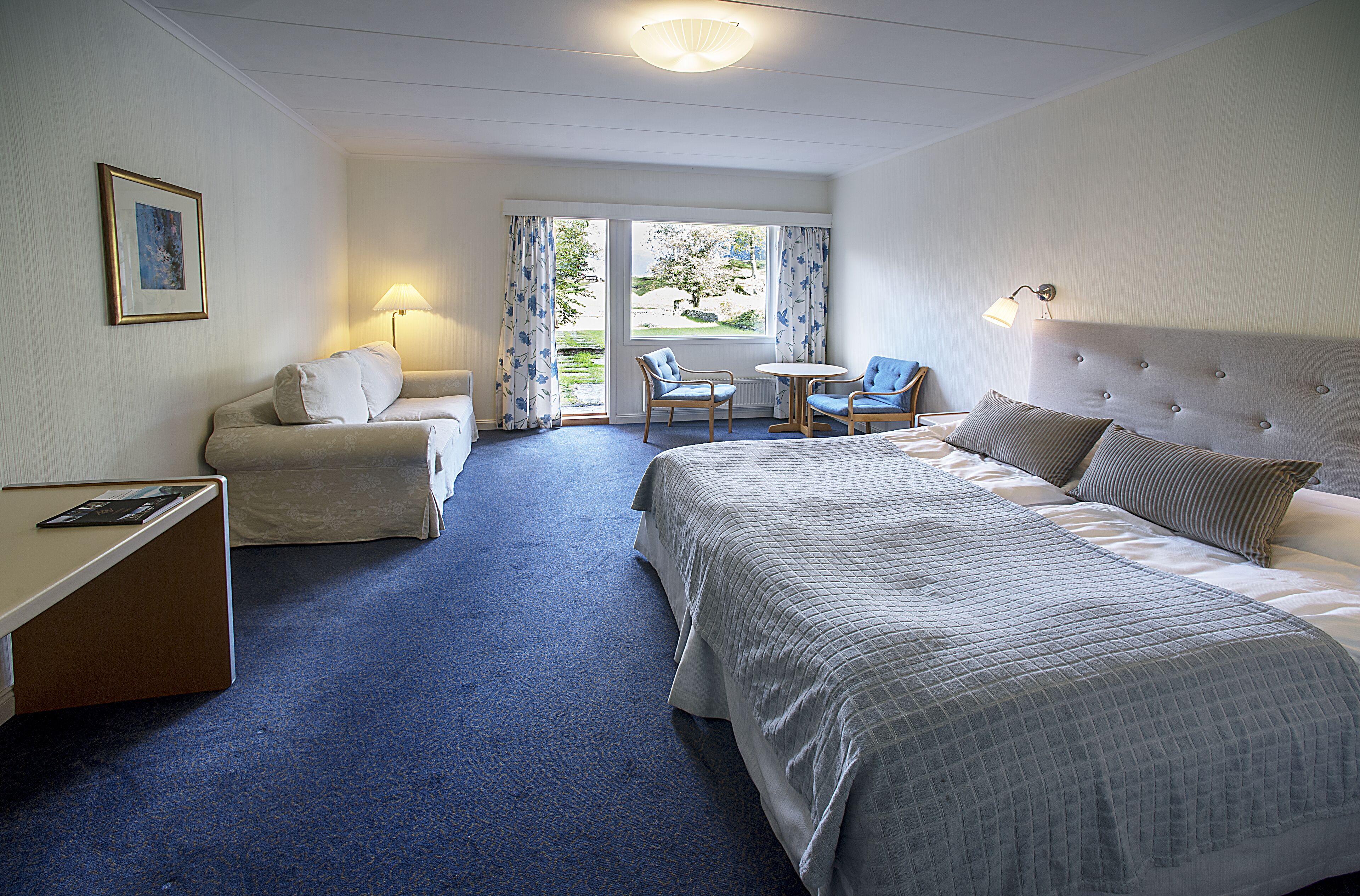 A hotel room with a bed, two armchairs, a small table, a desk, a lamp, and a window with curtains.