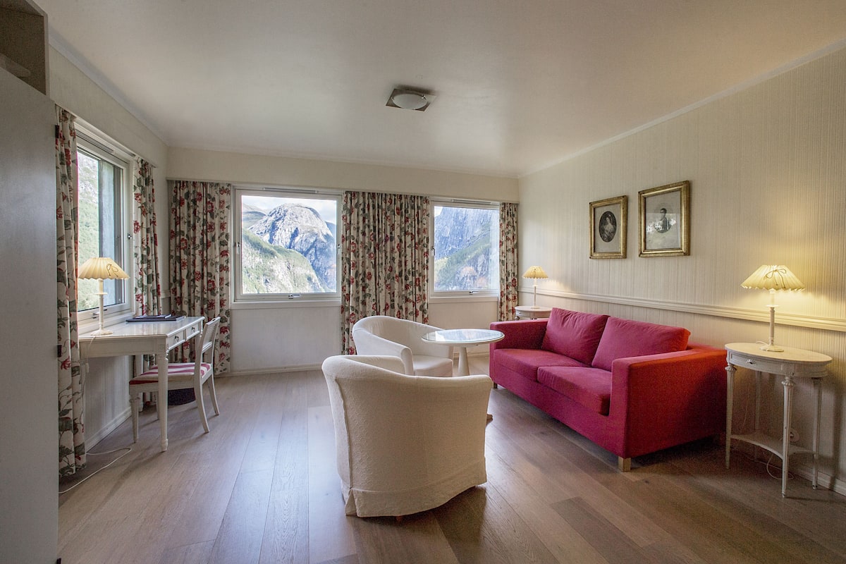 A room with a wooden floor, a red sofa, a white armchair, a small round table, and two windows with curtains.