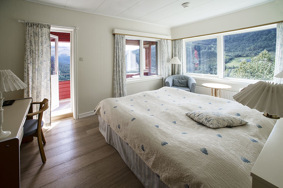 A bedroom with a large bed, a chair, a small table, and a view of the outdoors through a window.