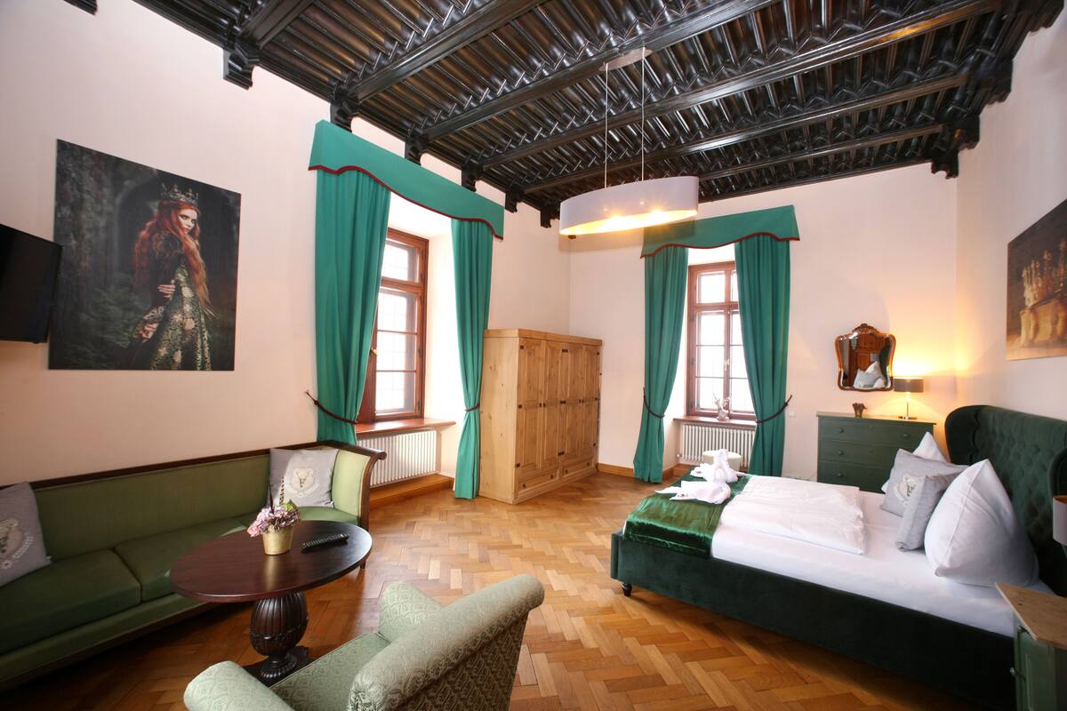 A room with a green sofa, a wooden cabinet, a bed with white bedding, and a green armchair.