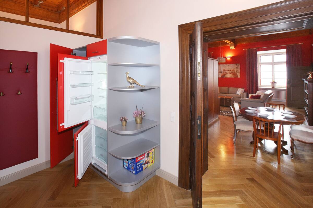 A compact kitchen with a built-in fridge, a grey shelving unit with decorative items, and a dining area with a wooden table and chairs.