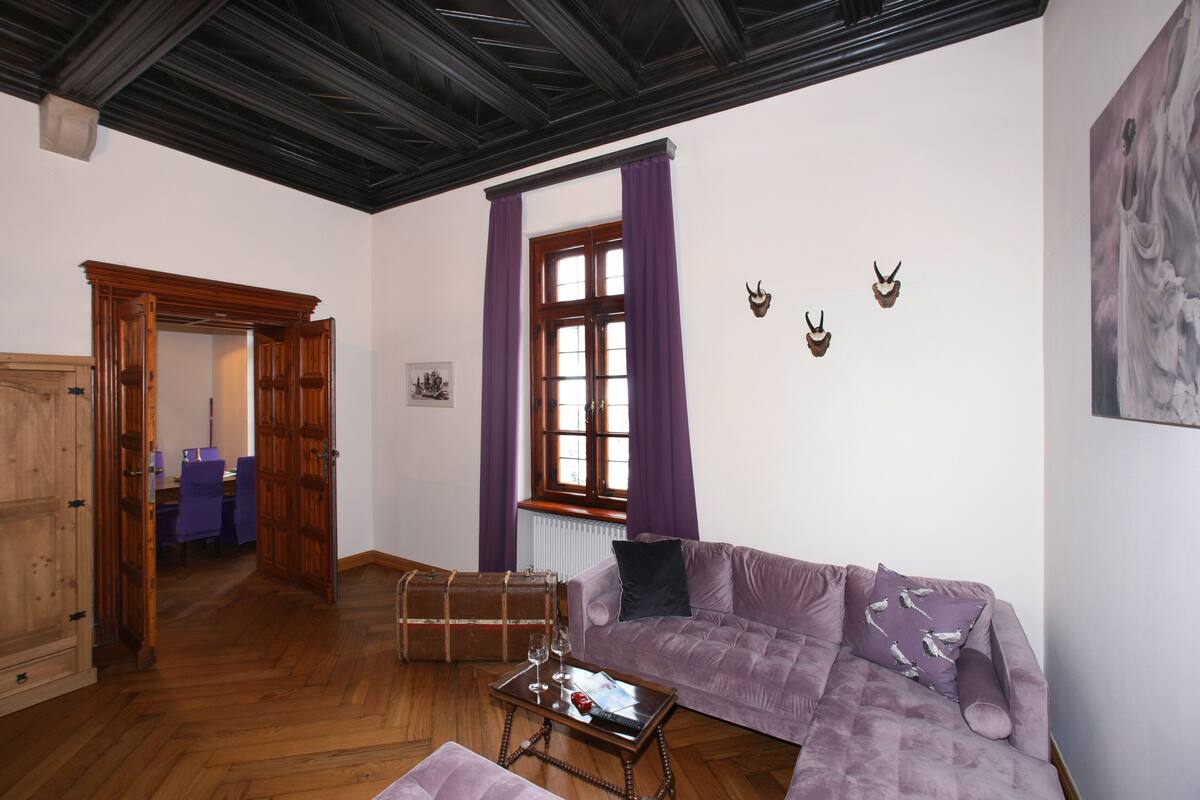 A room with a wooden floor, a purple sofa, a wooden table with a glass, and a window with purple curtains.