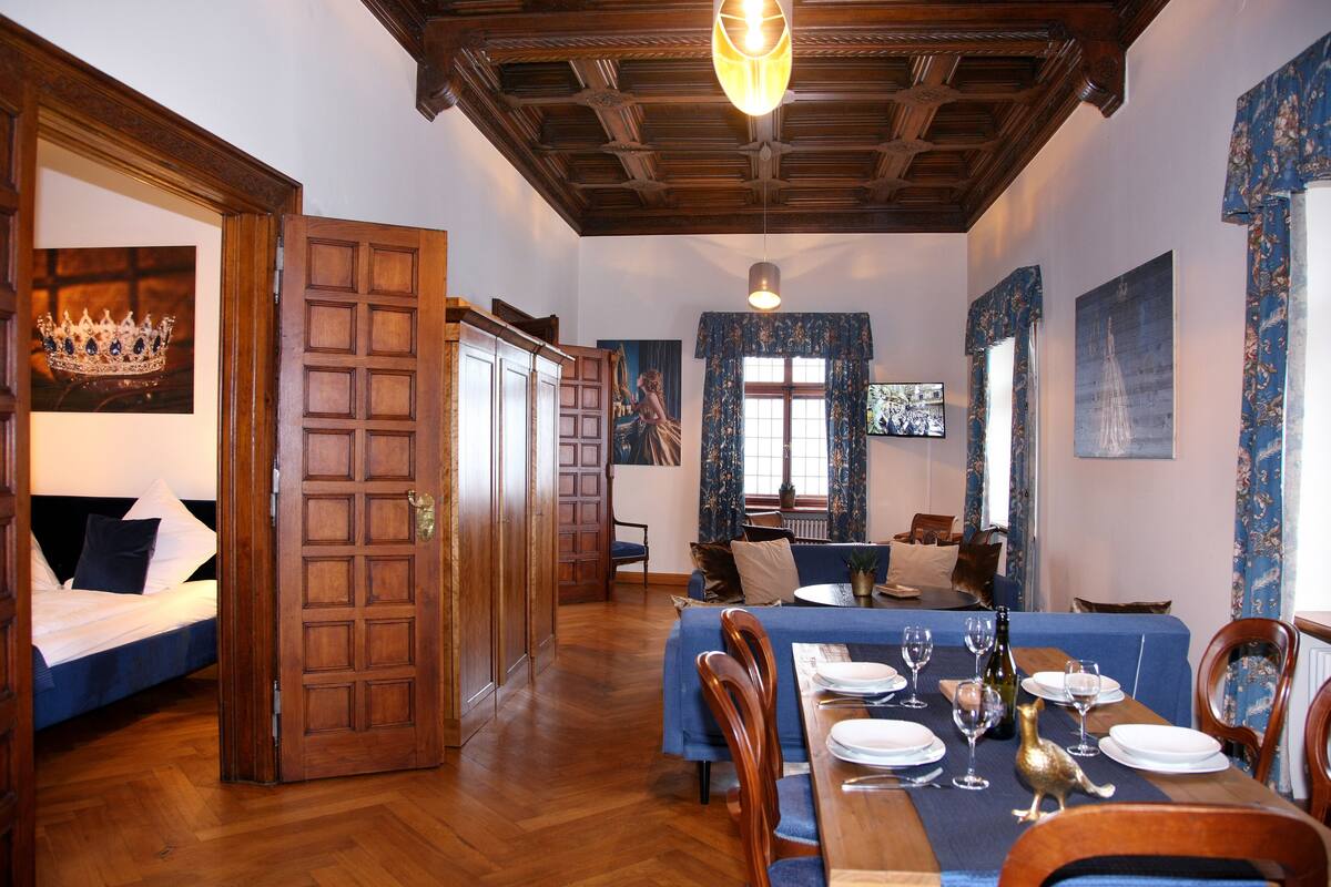 A dining area with a wooden table set for a meal, a wooden ceiling, and a large window with curtains.