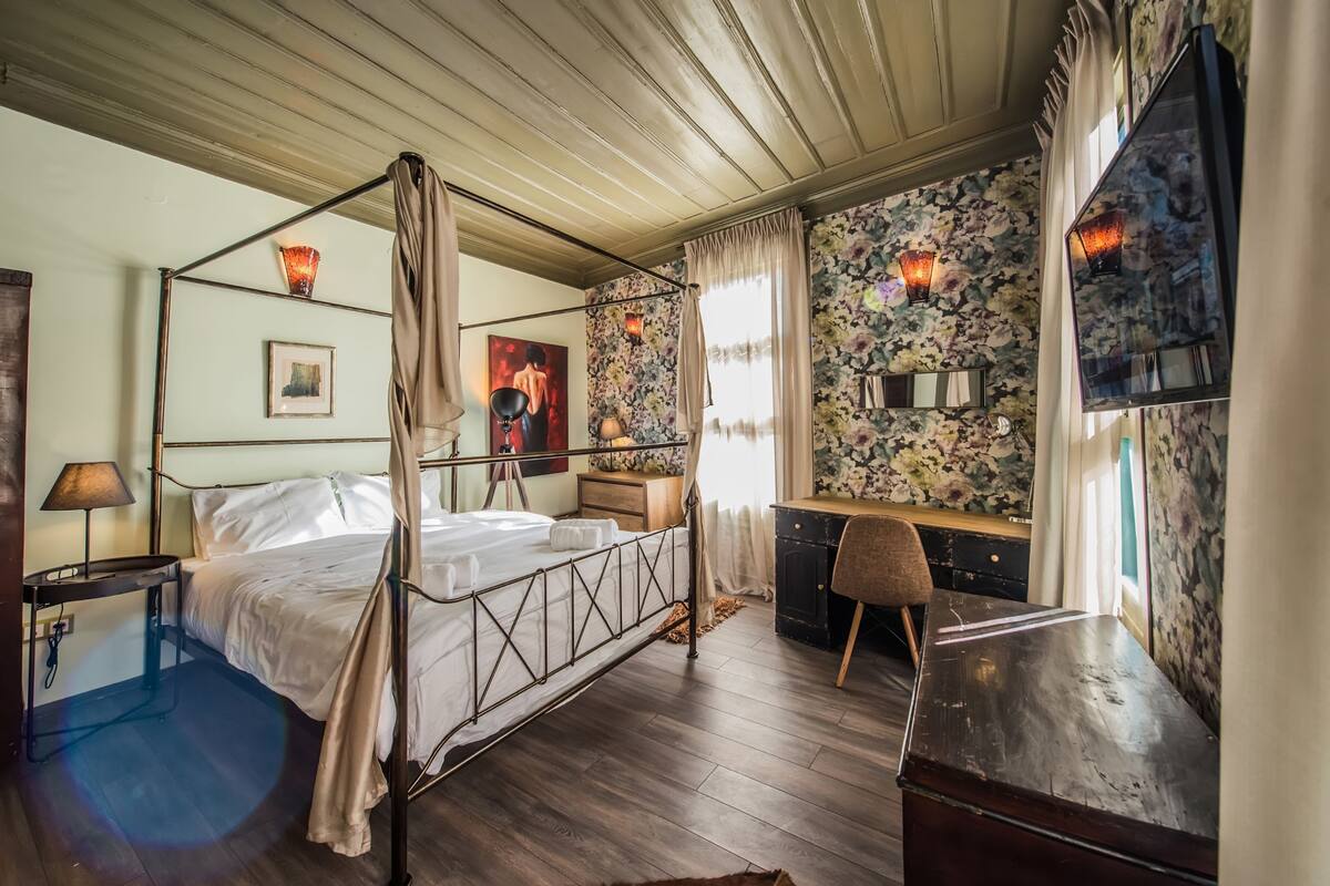 Un lit à baldaquin avec un plancher en bois, un bureau avec une chaise, une télévision et un mur recouvert d’un papier peint à motifs floraux.