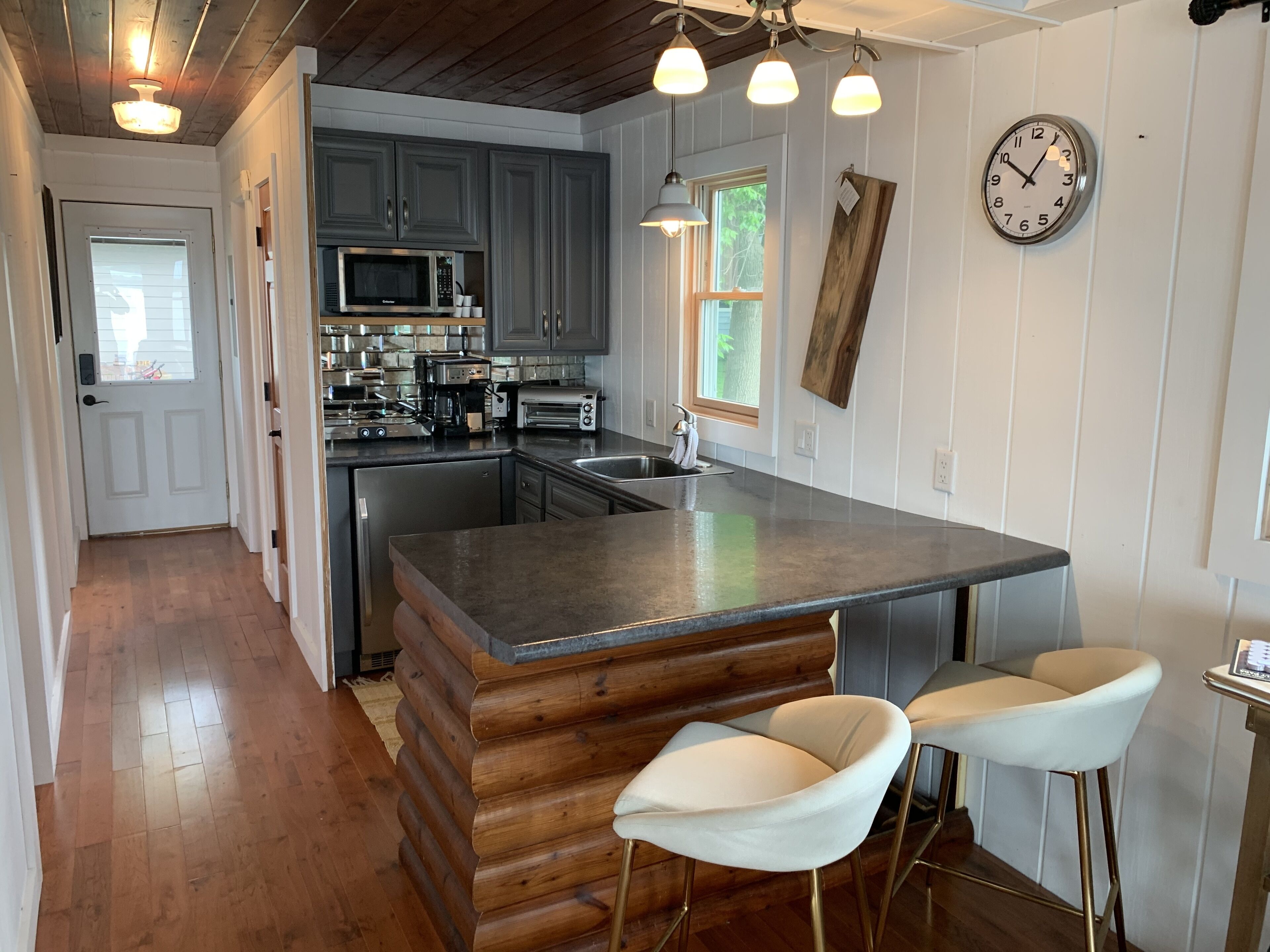 A modern kitchen with grey cabinets, a central island with bar stools, a microwave, and a clock on the wall.