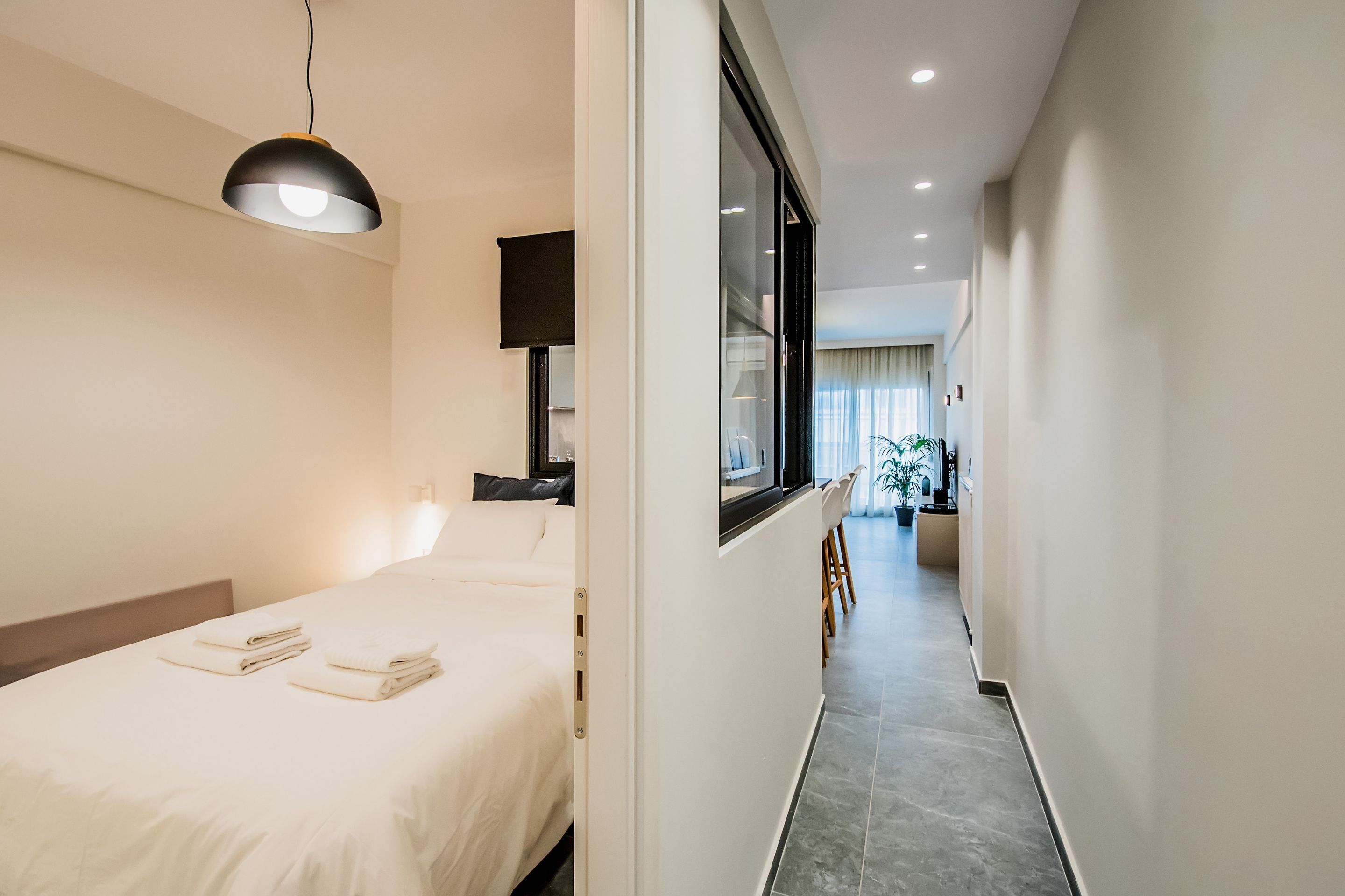 A hotel room with a bed, a desk, a chair, a window, and a hallway leading to another room.