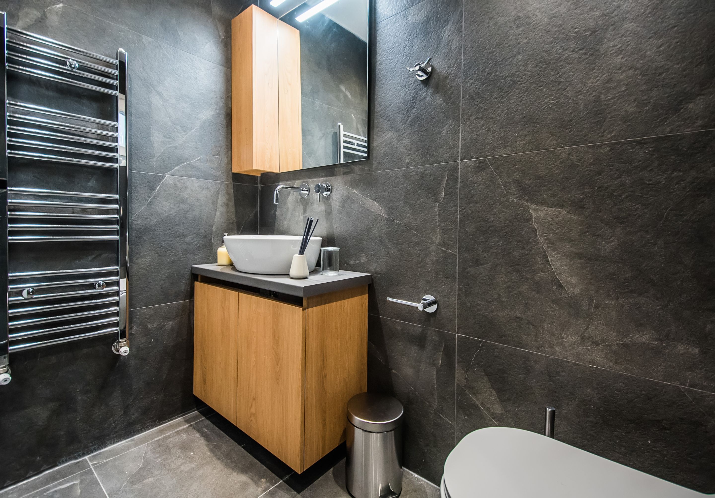 A modern bathroom with a wooden vanity, a white sink, and a stainless steel toilet.
