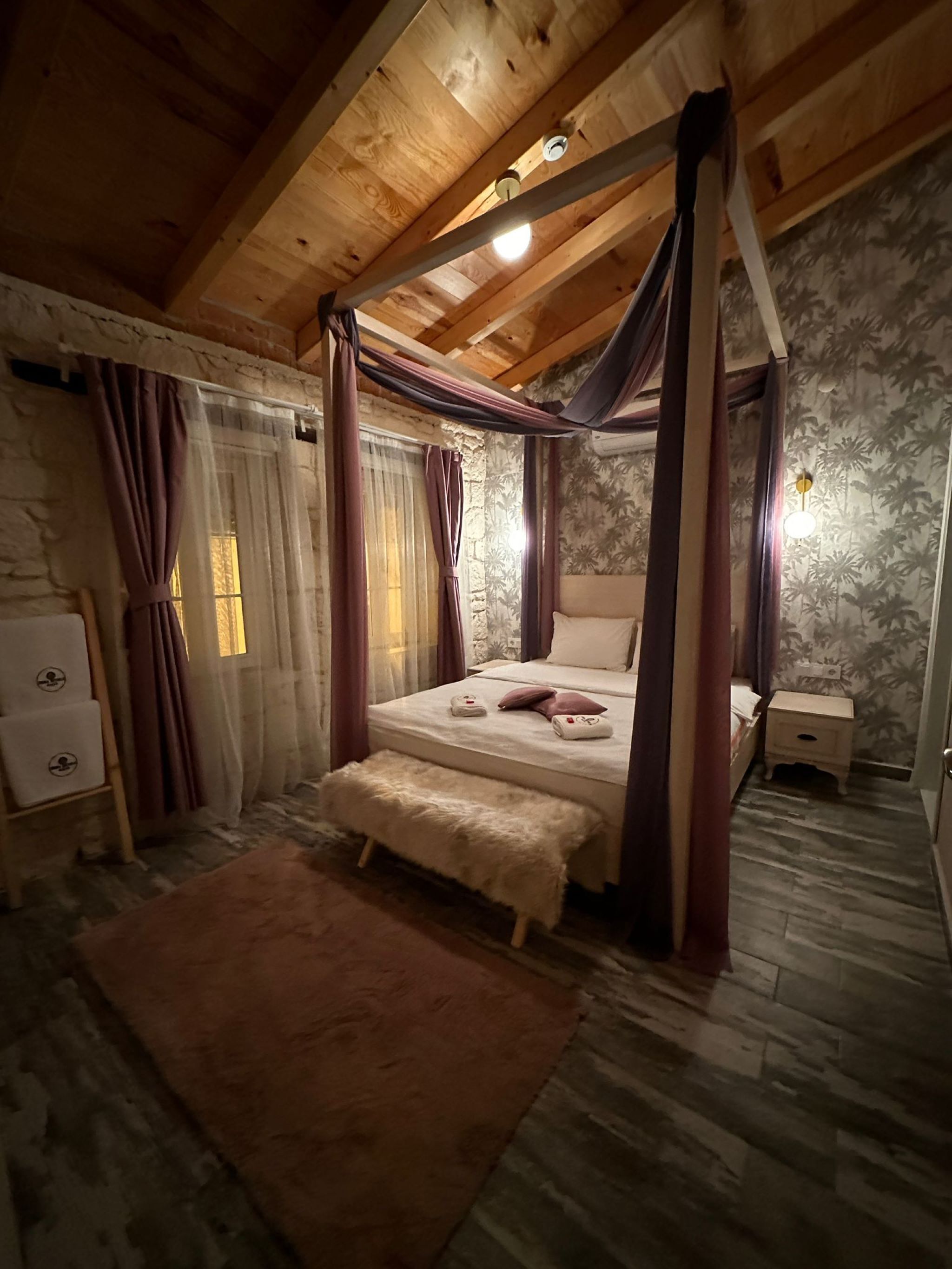 A room with a four-poster bed, a stone wall, a wooden ceiling, and a small bedside table.