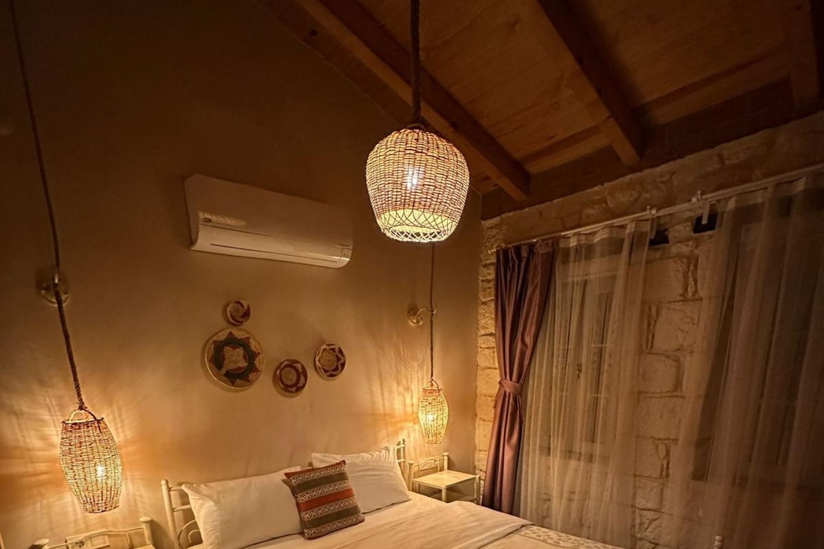 A cozy bedroom with a wooden ceiling, a bed with patterned bedding, two bedside lamps, and a stone wall.