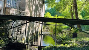 A rustic stone building with a metal bridge over a narrow water channel, surrounded by lush greenery and trees.