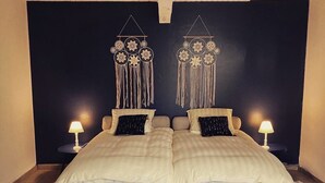 A double bed with a beige and white checkered duvet, two bedside tables with lamps, a round woven rug, and a dark wall with two macramé wall hangings.