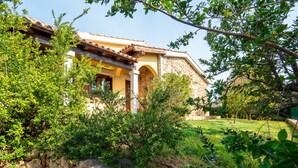 A traditional stone building with a tiled roof, surrounded by lush greenery and a stone wall.