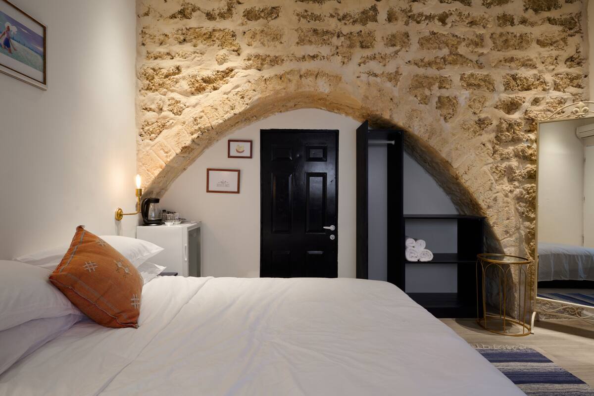 A bedroom with a stone archway, a bed with white bedding, a nightstand with a coffee maker, and a shelf with towels.