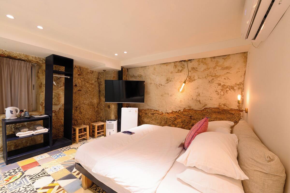 A bedroom with a large bed, a flat-screen TV mounted on the wall, a small bedside table, a shelf with towels, and a patterned floor.