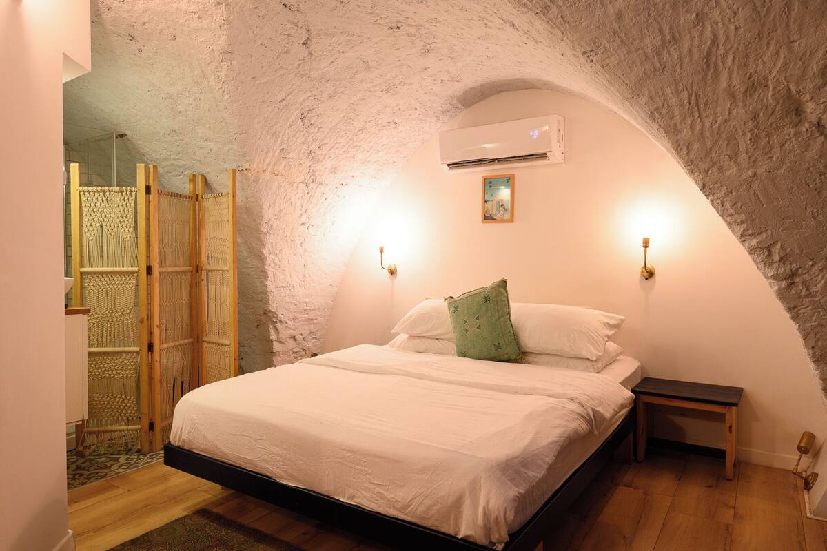 A bedroom with a stone ceiling, a bed with white linens, two wall-mounted lights, and a small bedside table.