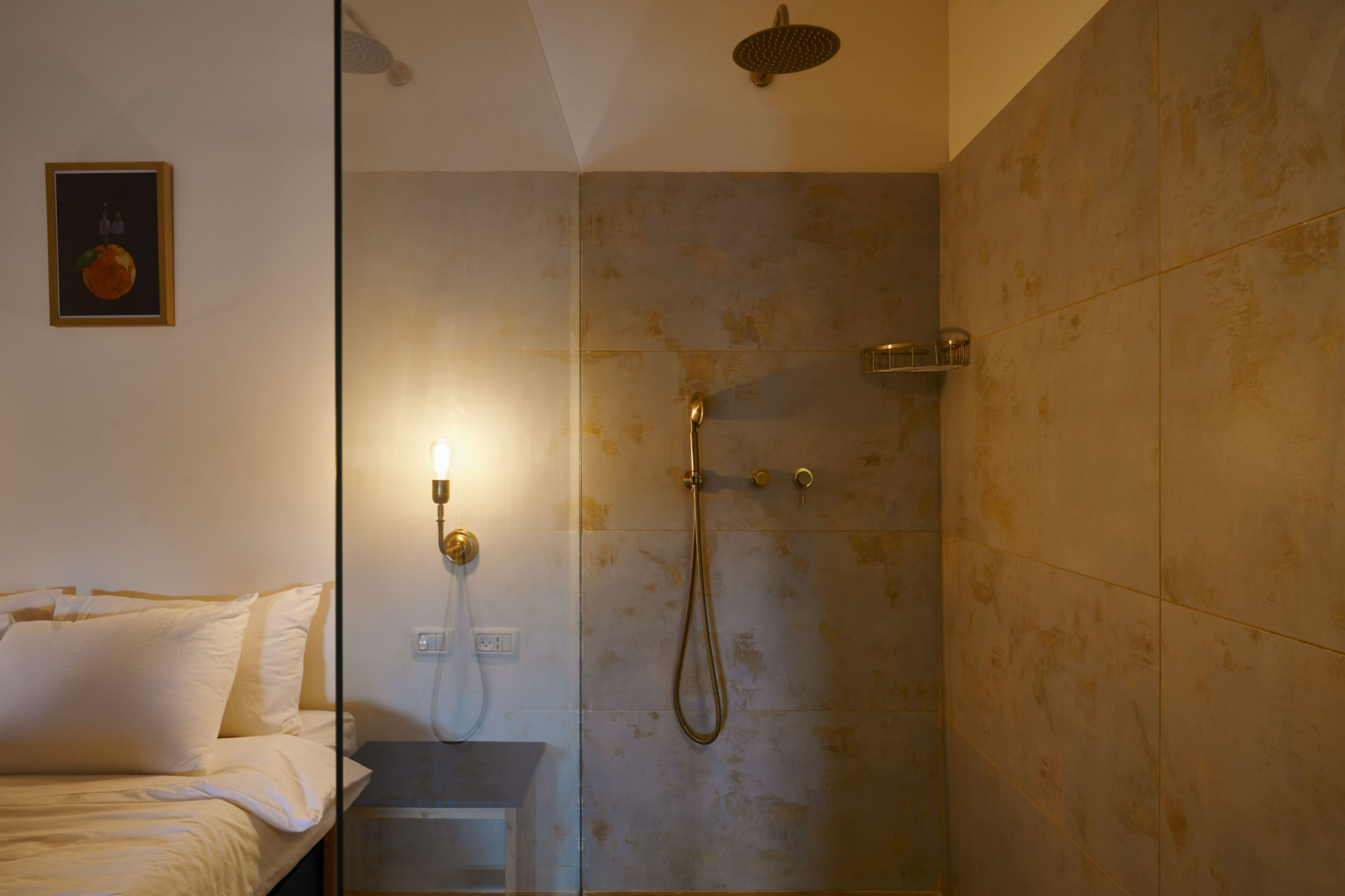 Habitación de hotel con cama, mesita de noche, lámpara de pared y zona de ducha con cabezal de ducha de mano y grifo de pared.
