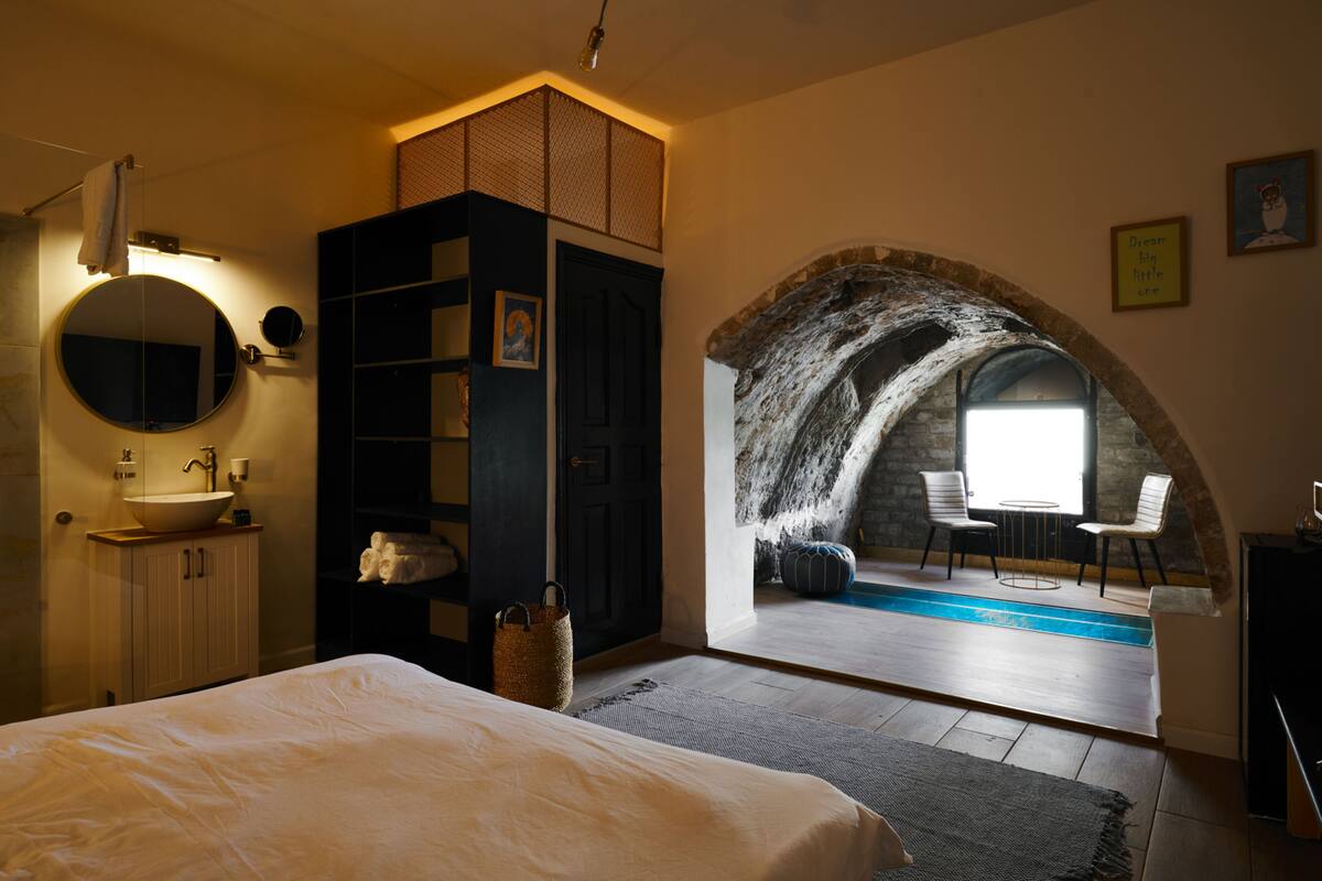 A bedroom with a large bed, a round mirror, a dark wardrobe, a small bathroom with a sink, and a view of an outdoor swimming pool through an arched stone window.