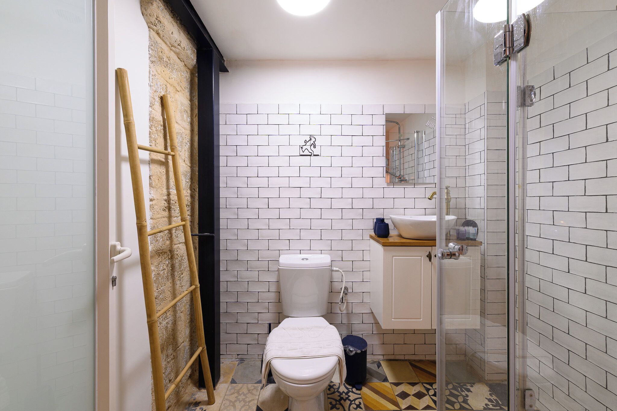 Un baño pequeño con inodoro, lavabo y ducha. Una escalera está apoyada en la pared, y se ve un cubo de basura.