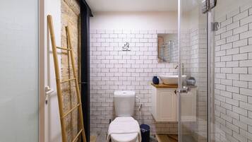 Un baño pequeño con inodoro, lavabo y ducha. Una escalera está apoyada en la pared, y se ve un cubo de basura.