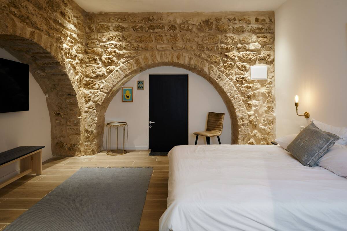 A bedroom with a stone archway, a bed, a chair, and a television mounted on the wall.