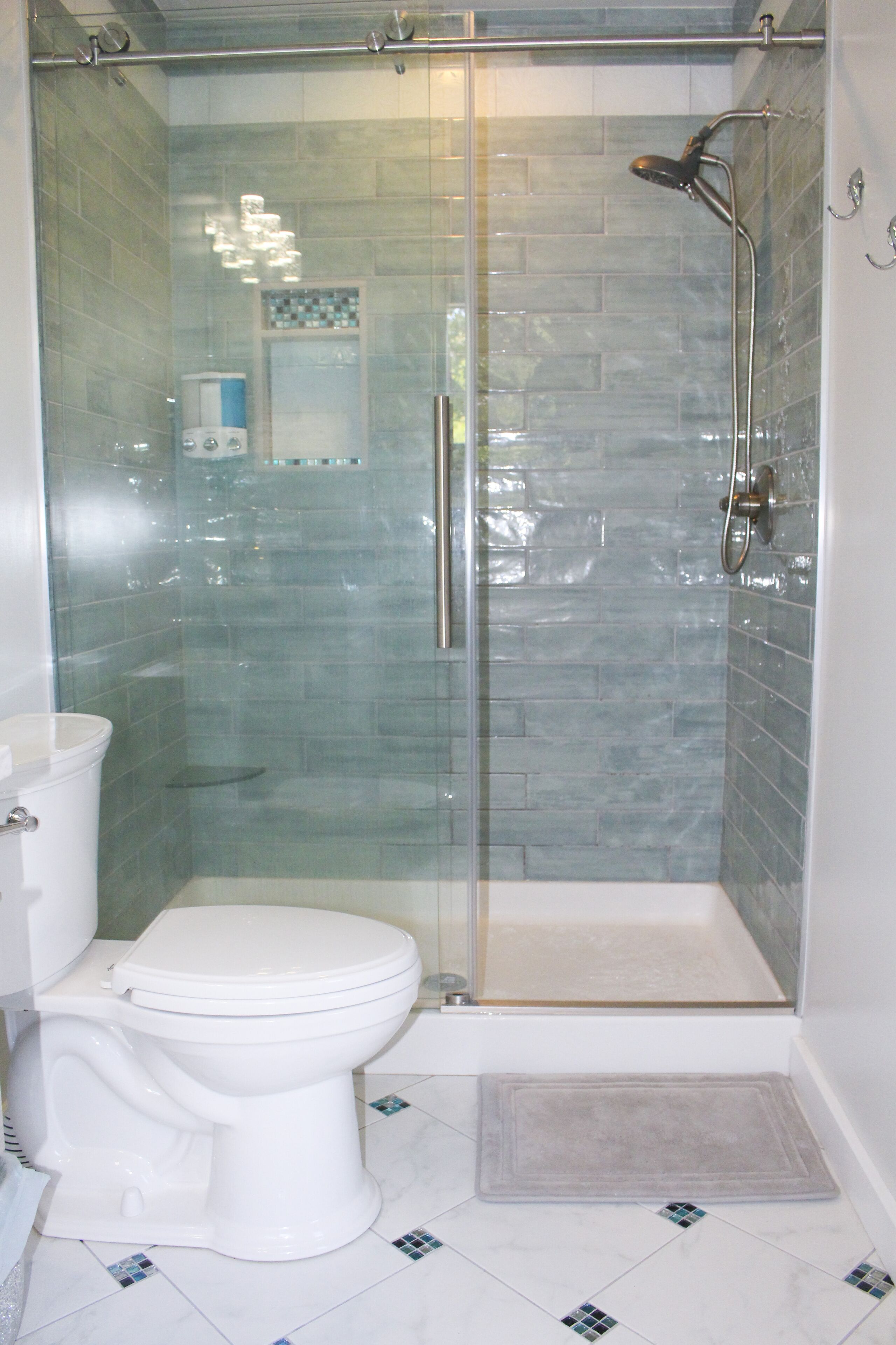 A glass-enclosed shower with a handheld showerhead, a white toilet, and a small rug on the floor.