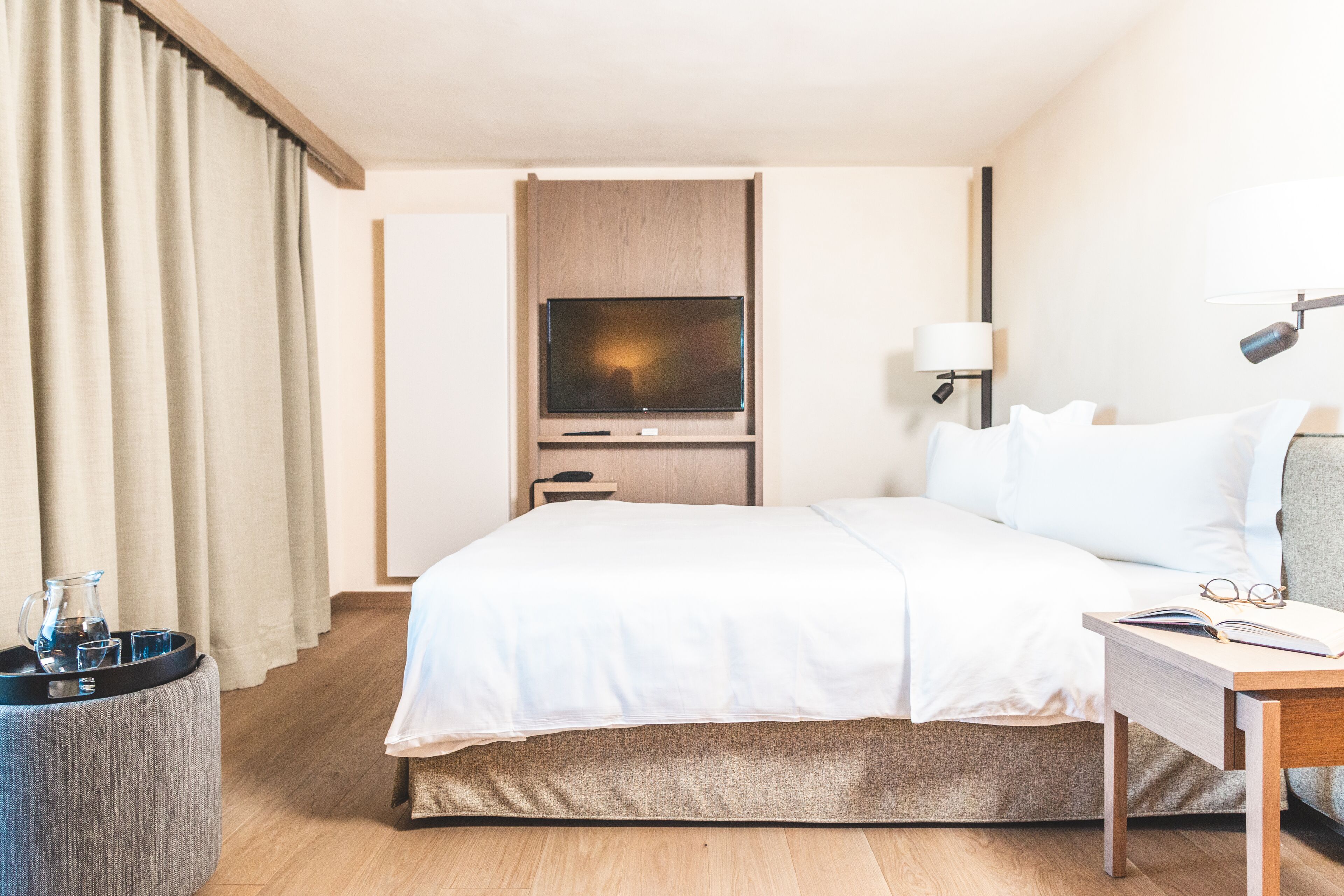 A hotel room with a bed, a TV, a bedside table with glasses, a lamp, and a small table with a pitcher and glasses.
