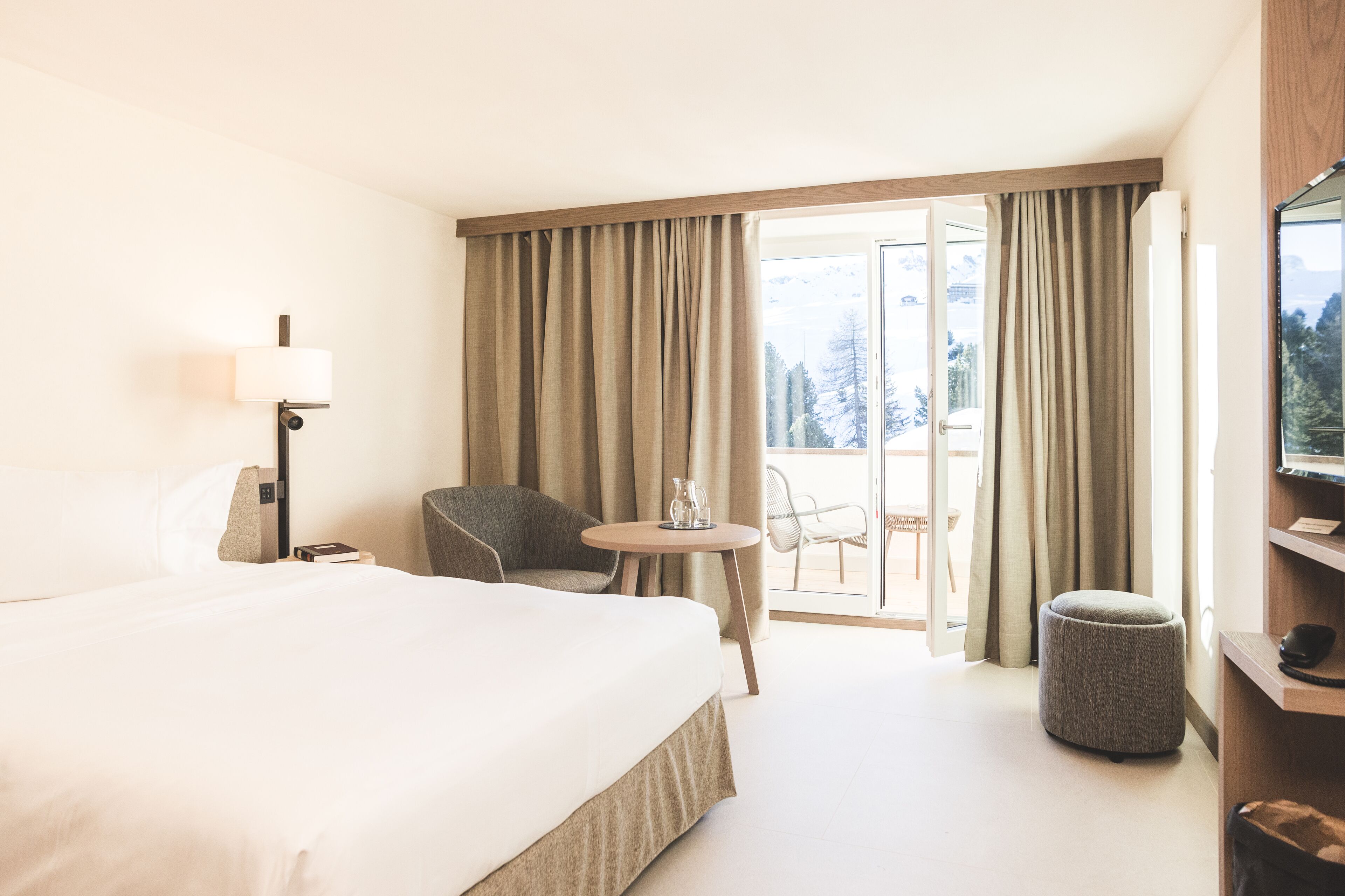 Quarto de hotel com uma cama grande, uma escrivaninha com abajur, uma cadeira, um banquinho redondo pequeno e uma varanda com cortinas.