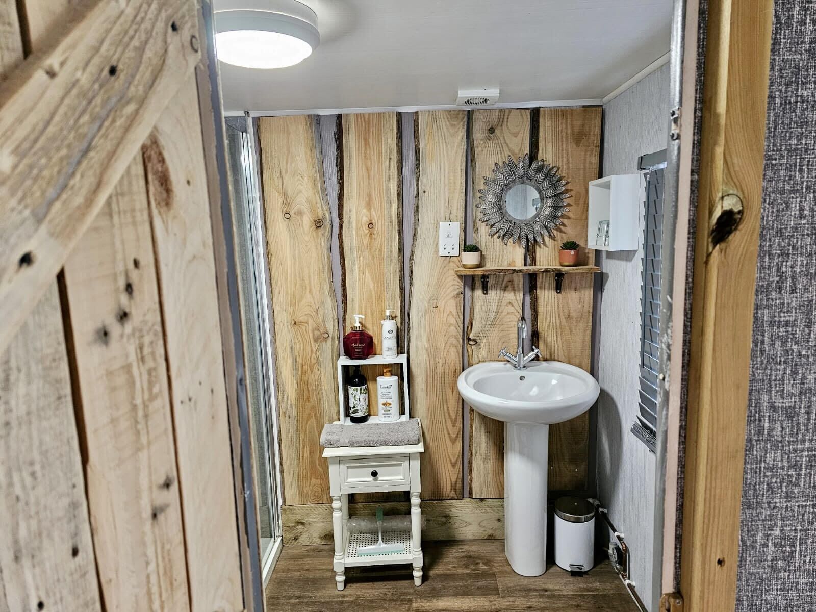 Un baño con panel de pared de madera, un lavamanos pedestal blanco, un tocador pequeño blanco con espejo y un estante con artículos de aseo.