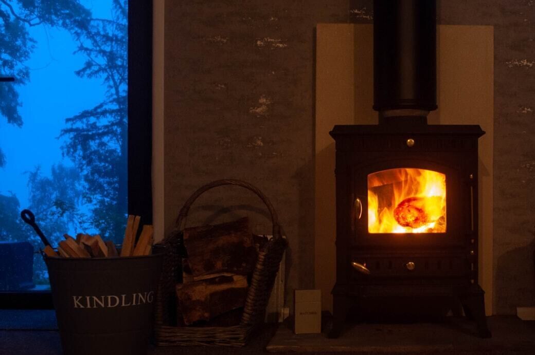 A wood-burning stove with logs in a basket labeled "KINDLING" and a window with a view of trees.