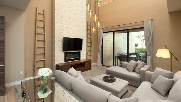 A modern living room with a flat-screen TV, beige sofas, a wooden entertainment unit, and a large window with a view of the outdoors.