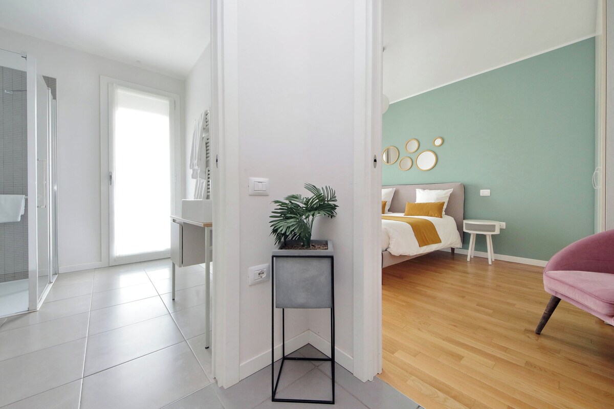 A modern apartment with a bathroom, a small plant on a side table, a bed with a headboard, and a pink chair.