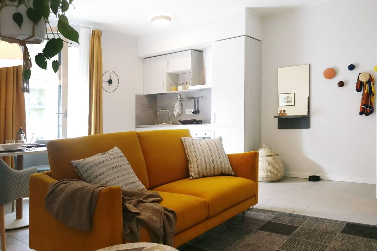 Uma sala de estar moderna com um sofá amarelo mostarda, uma mesa de centro redonda e uma cozinha ao fundo.