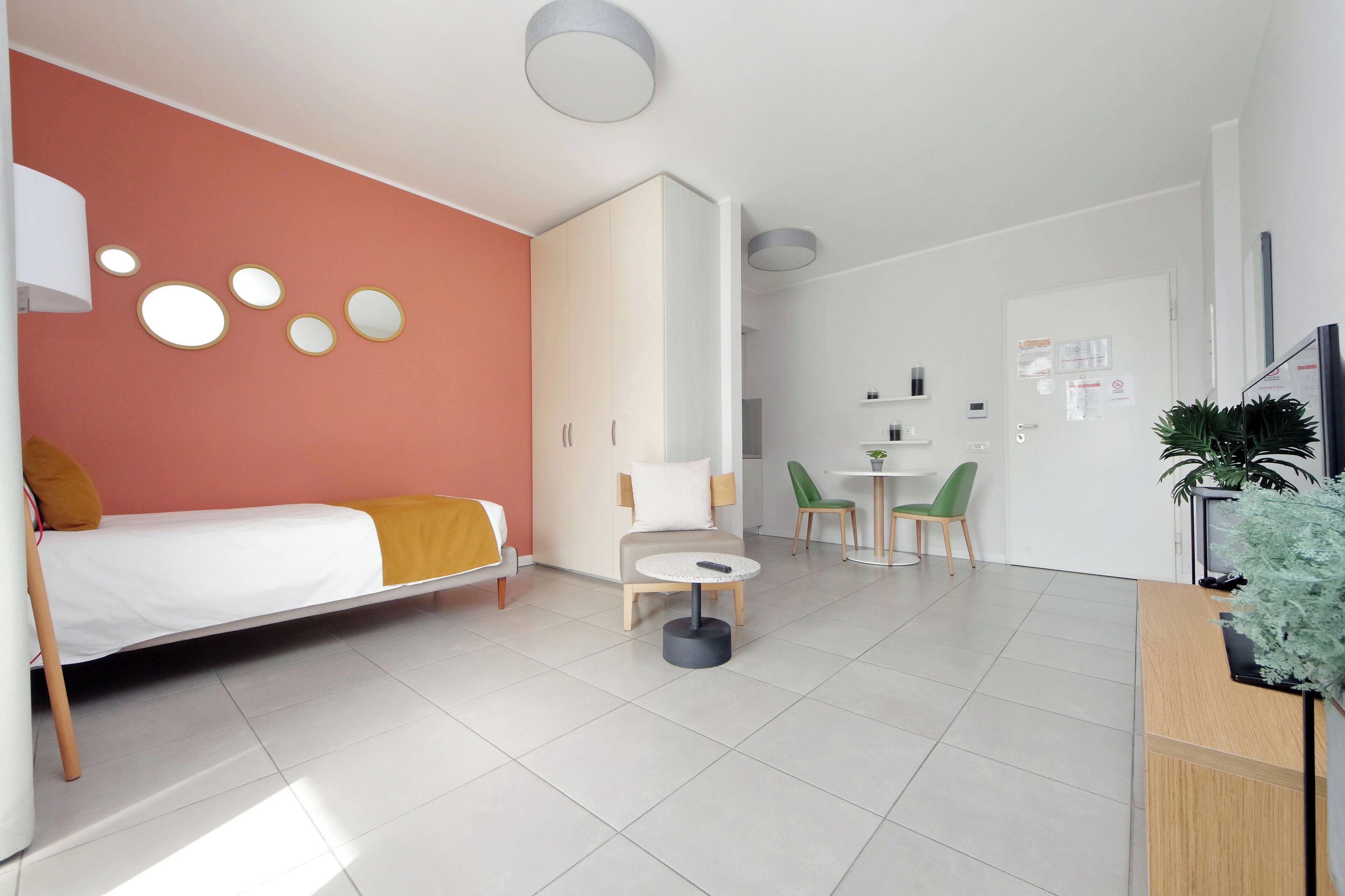A modern hotel room with a bed, a small table, a sofa, a desk, and a reception area.