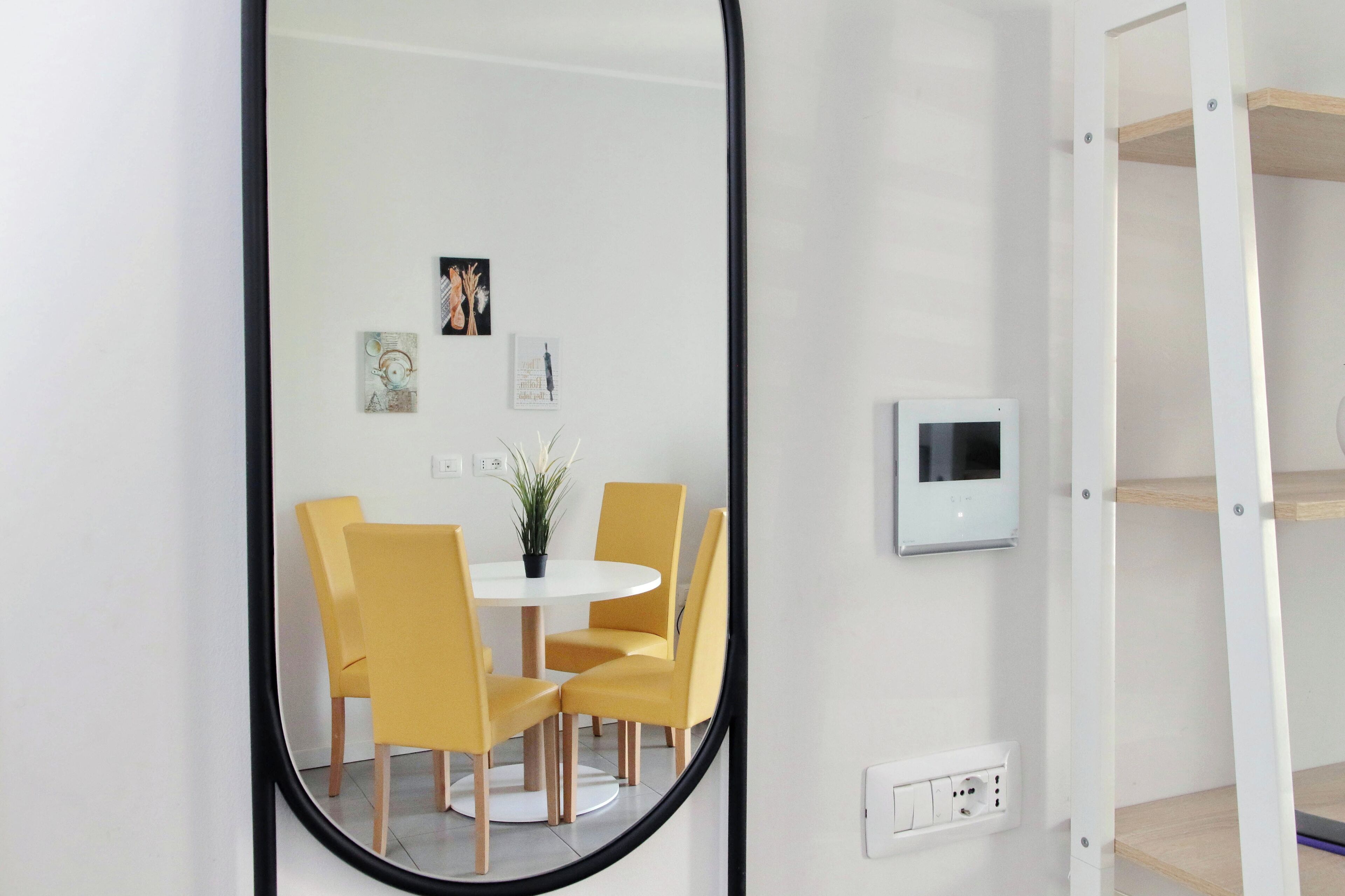 A room with a round table, four yellow chairs, a potted plant, and a white wall-mounted thermostat.