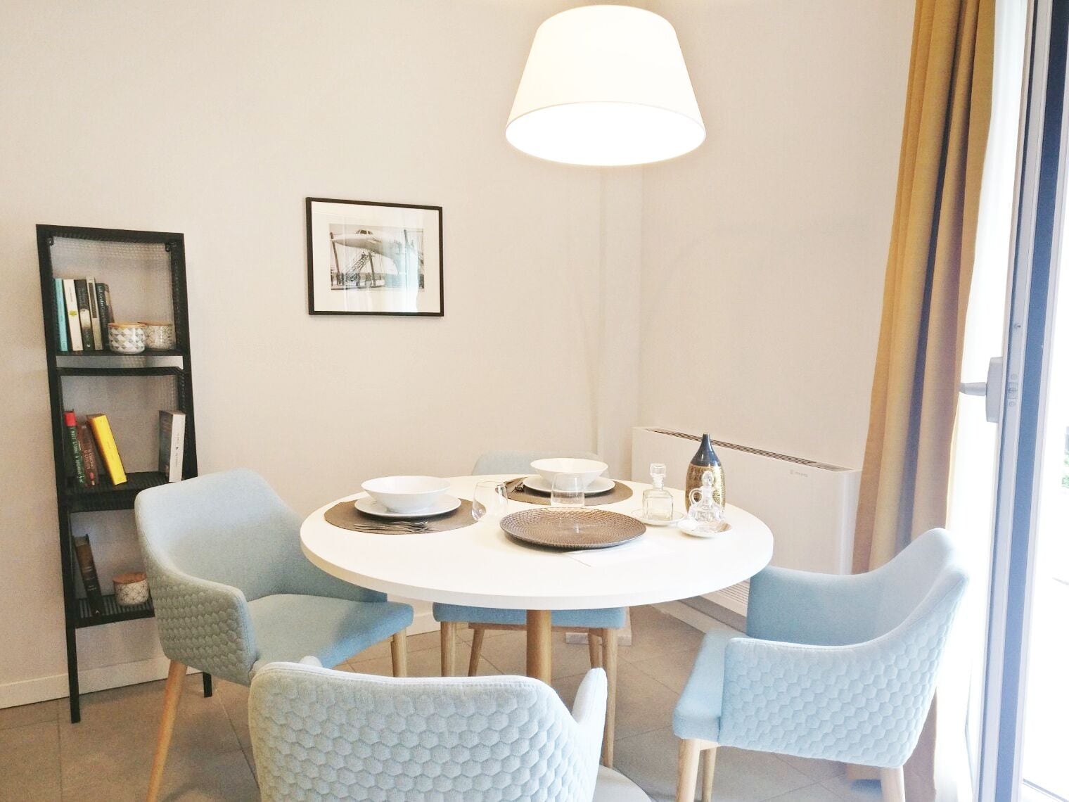 A dining area with a round table, four light blue chairs, a white tablecloth, and a hanging light fixture.