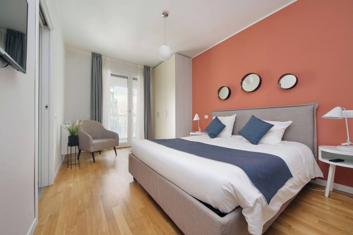 Um quarto moderno com uma cama grande, uma cadeira, uma mesinha de cabeceira e uma janela com cortinas.