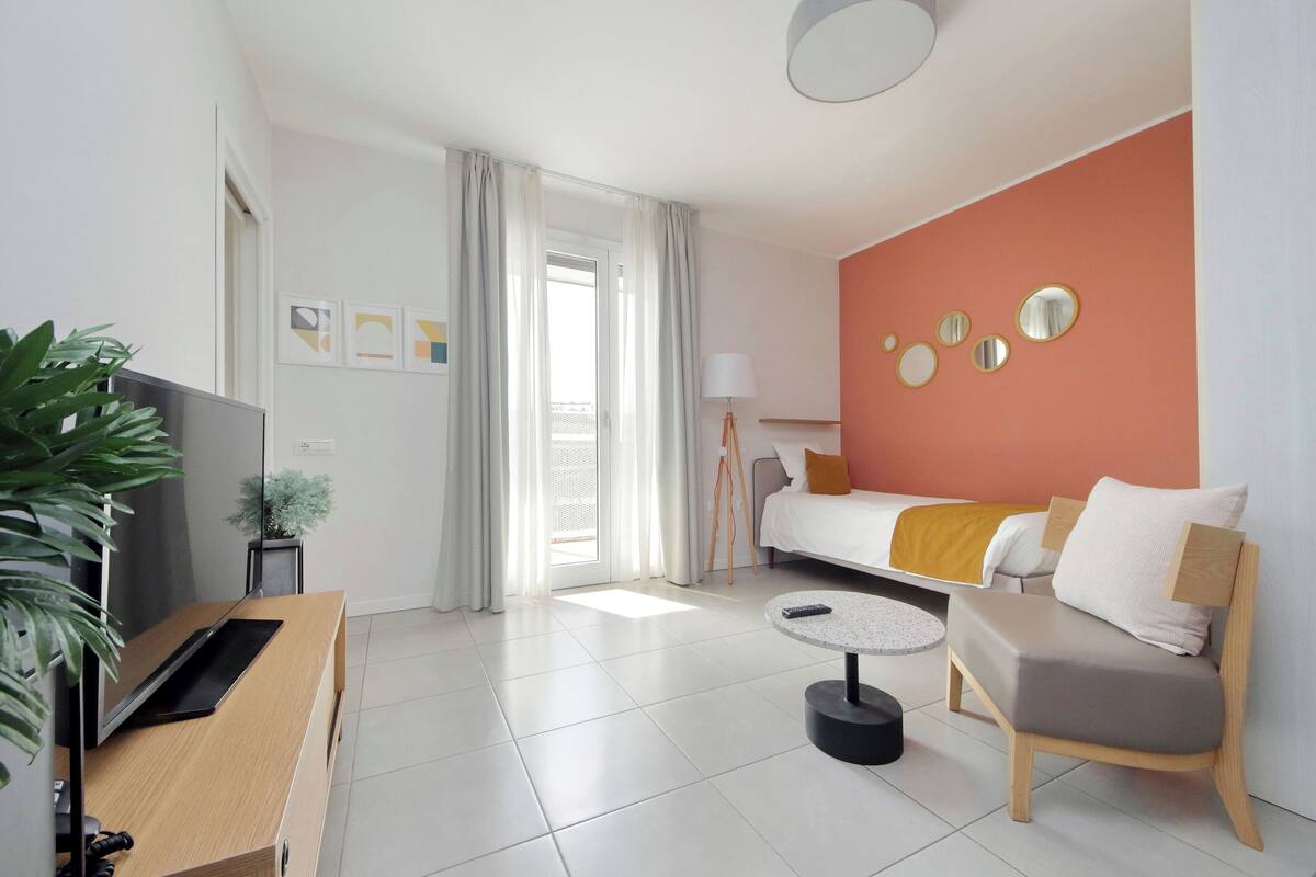 Quarto de hotel moderno com sofá, uma mesa redonda pequena, televisão e um vaso de planta.