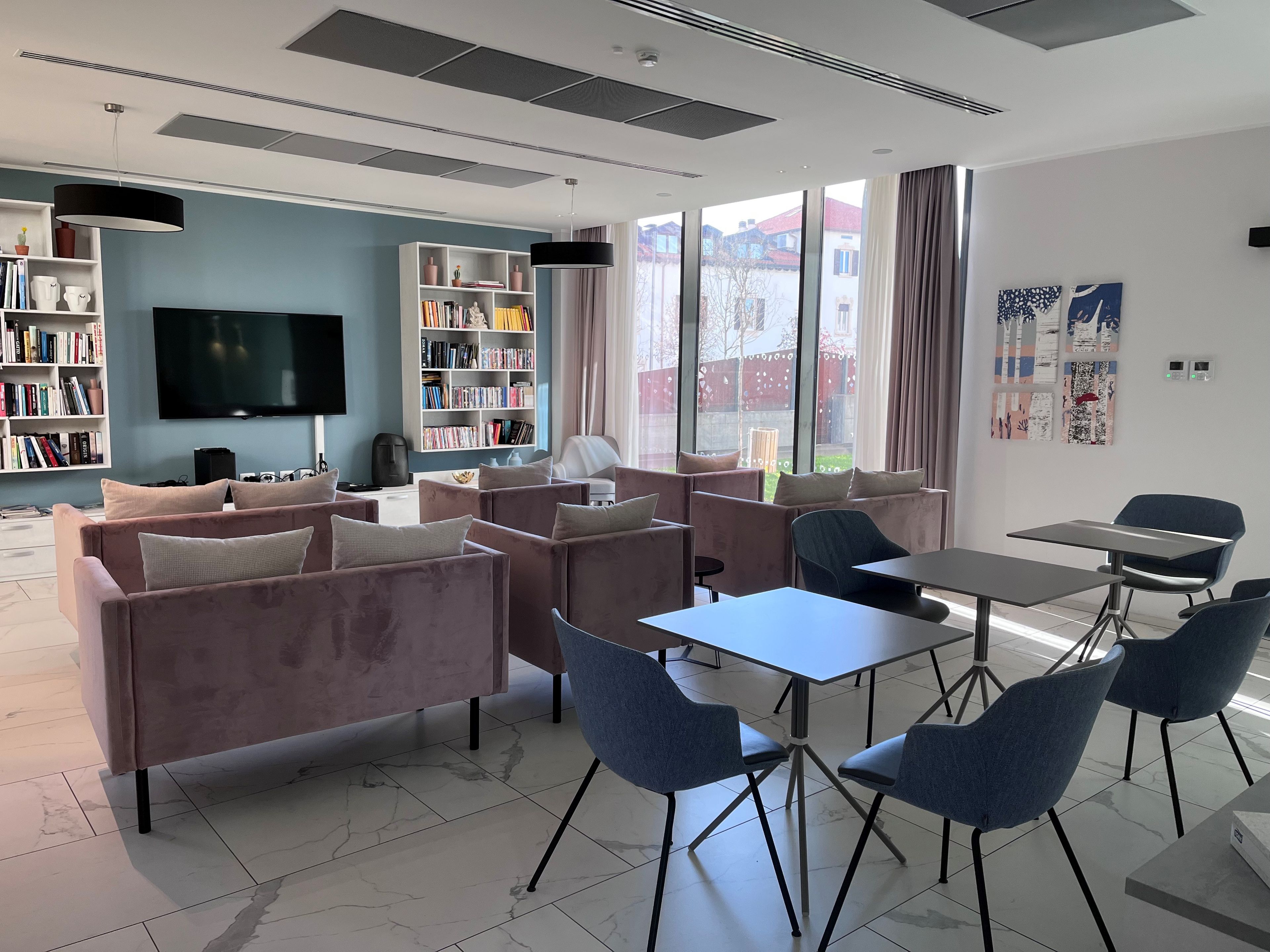 A modern living room with sofas, a coffee table, and a TV. There are bookshelves and a large window with a view.