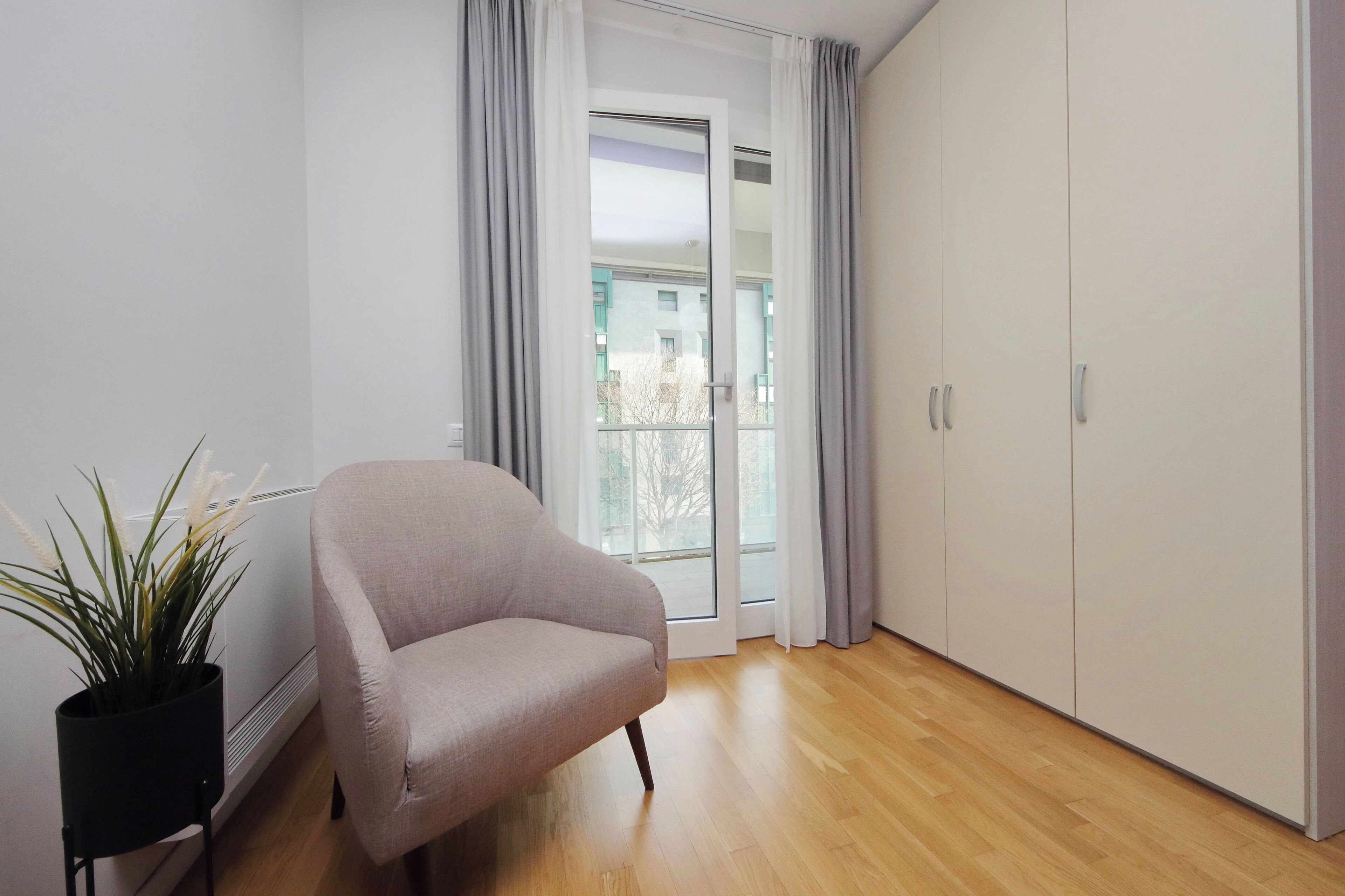 A room with a grey armchair, a potted plant, a wardrobe, and a sliding glass door leading to an outdoor area.