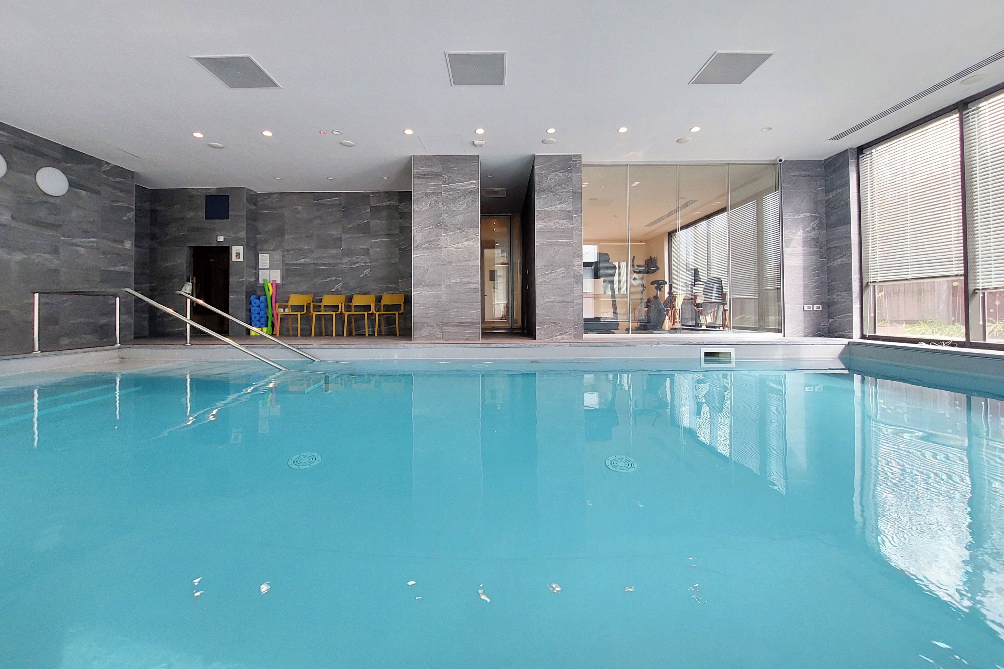 Ein Hallenbad mit freiem Blick auf den Poolbereich, ein Sitzbereich mit Stühlen und eine Glasswand, die in einen anderen Raum führt.
